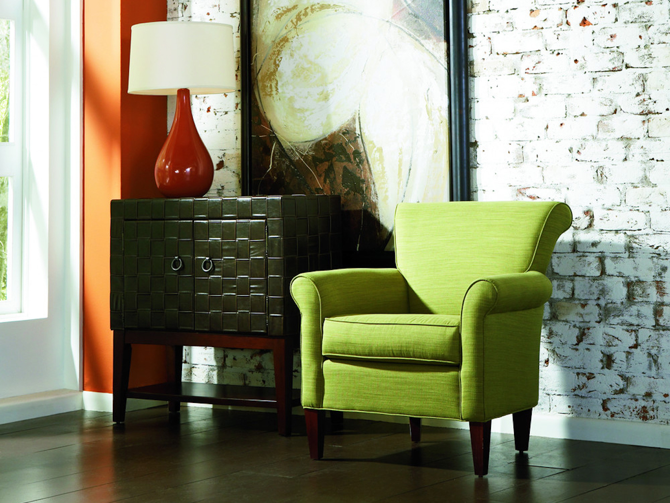 Green armchair, woven black chest with a red table lamp in front of an exposed brick wall