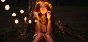 Girl with red hair sitting in front of a tree, holding white string lights