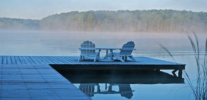 Empty Adirondack chairs and table sitting on a dock overlooking misty lake with trees in background