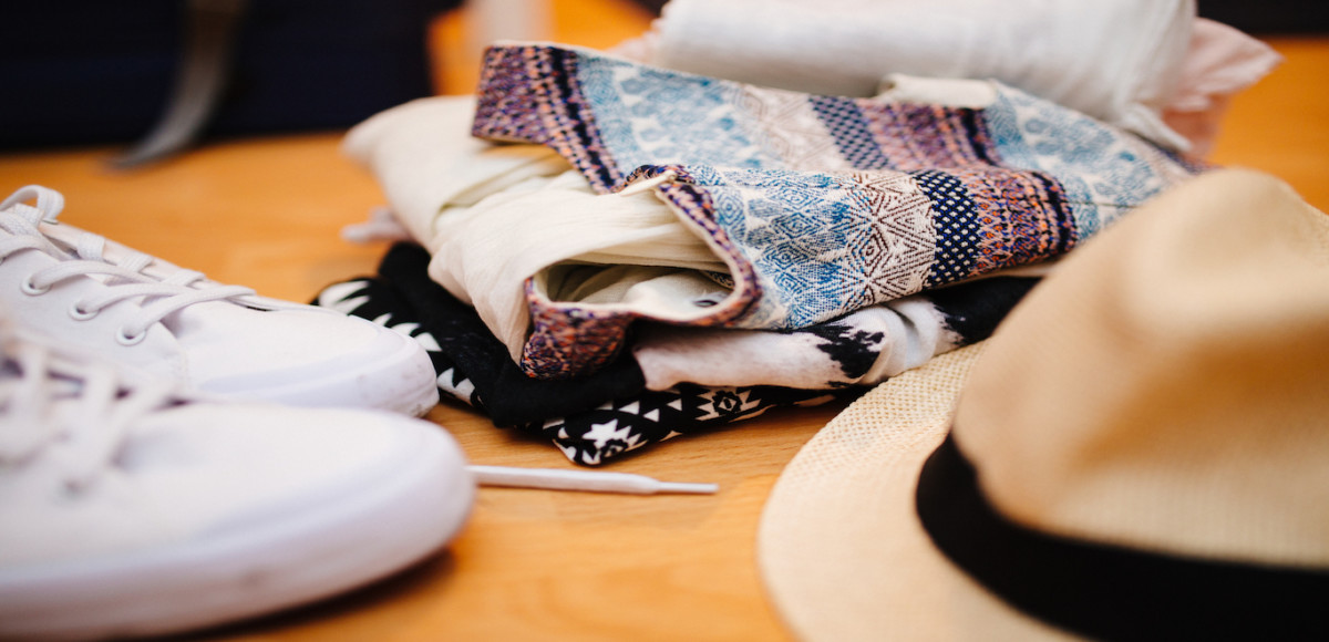 Close-up of white tennis shoes, fedora hat, and colorful folded clothes on wood floor ready for packing