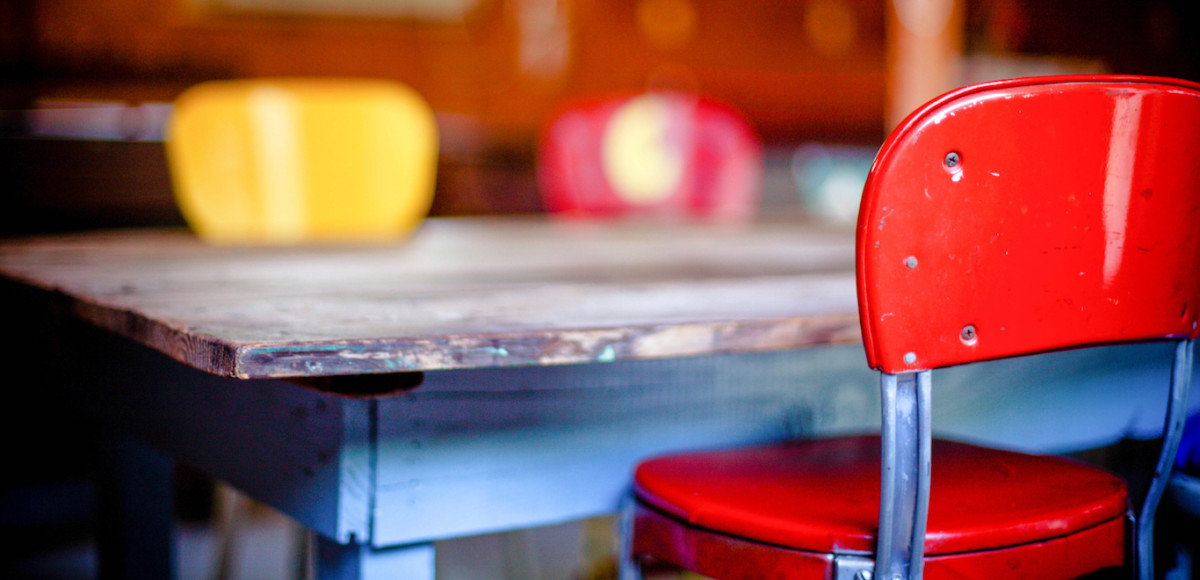 Close-up of a vintage red chair and scuffed wooden table with yellow and red chairs in the blurred background