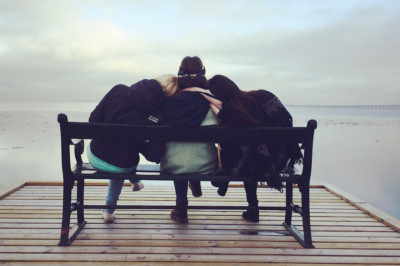 Two friends rest their heads on the shoulders of a third friend while sitting on a bench on a dock overlooking a lake