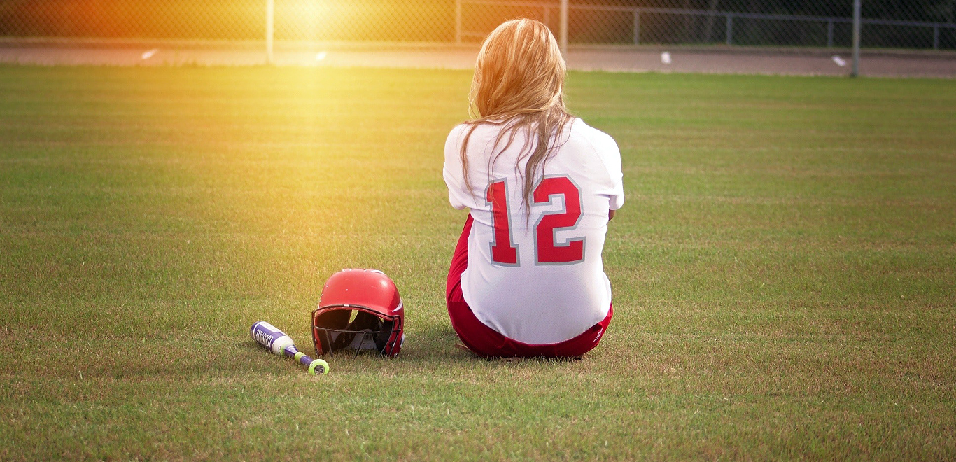 Woman in baseball jersey sitting in field with bat and helmet at sunset.