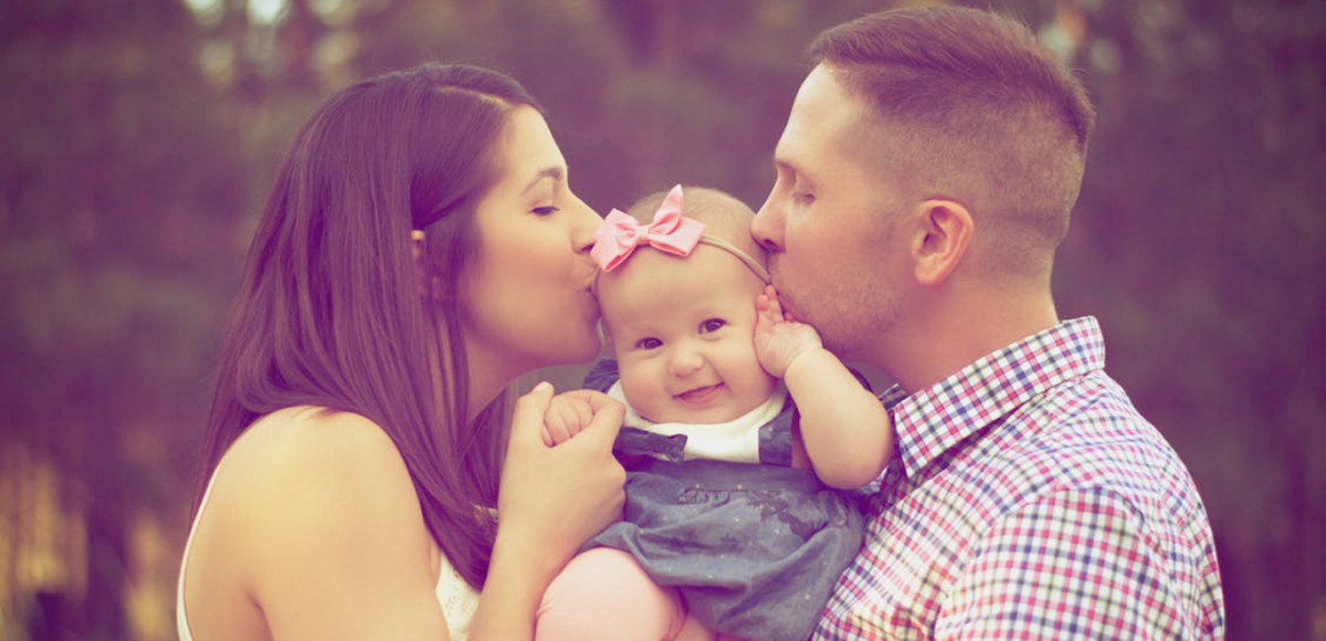 Mom and dad kissing a smiling baby outdoors