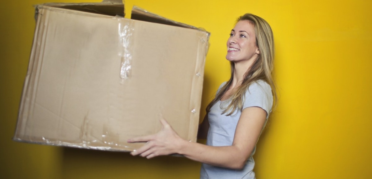 Smiling woman holding a large brown moving box against a yellow background