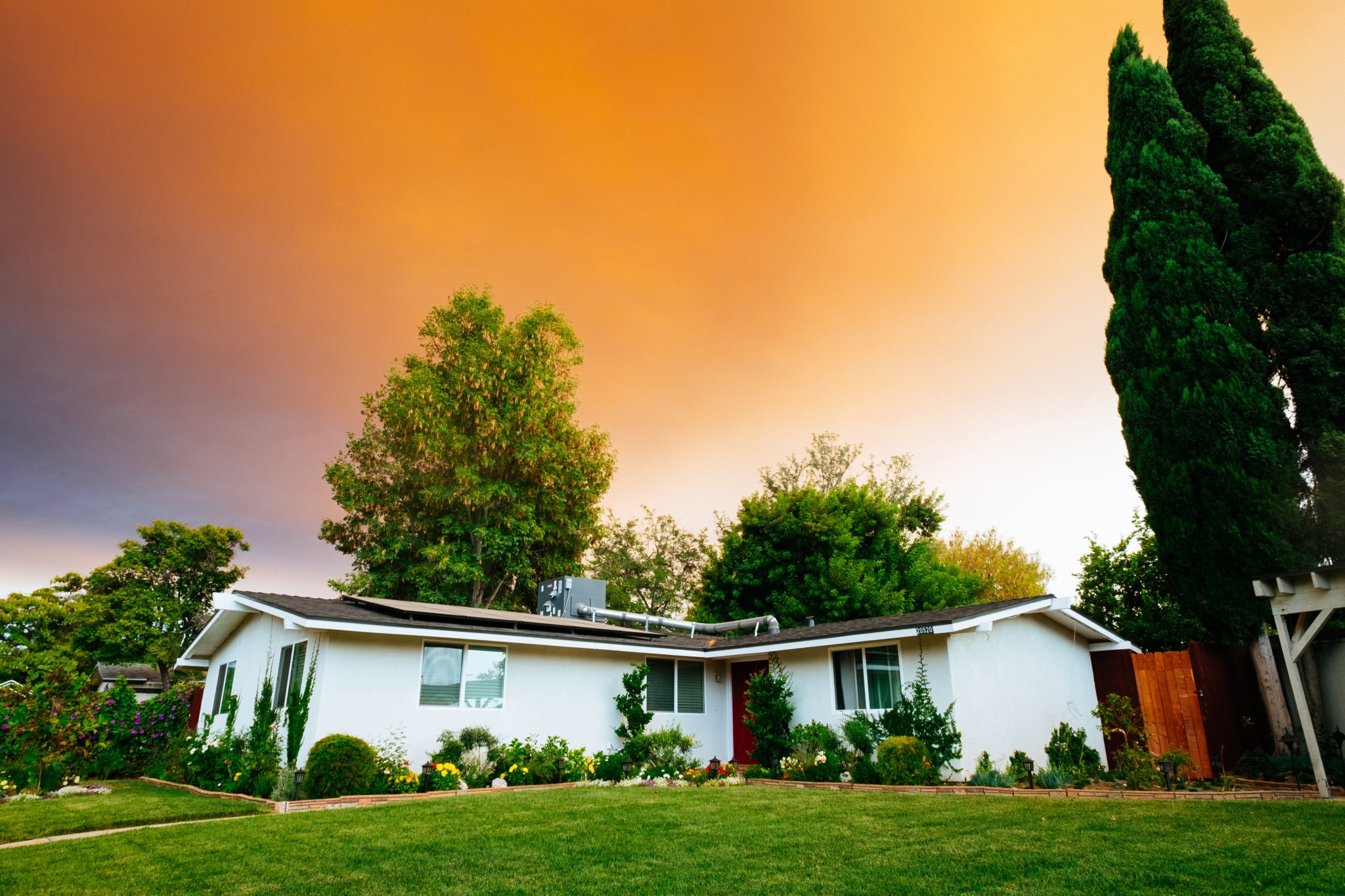 A small white house in a suburban neighborhood surrounded by green trees with an orange sunset in the background