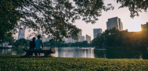 Two people sitting on a park bench overlooking a river with the city skyline with tall buildings in the background.