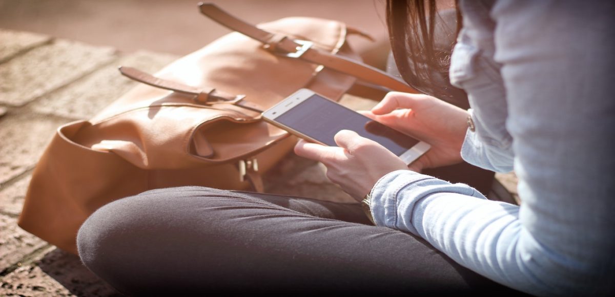 Young female college student sitting cross-legged outdoors using smartphone, with satchel nearby
