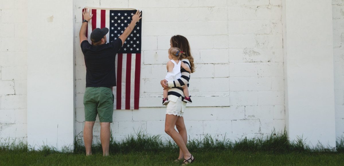 Man holding U.S. flag up against the exterior of a white building with woman holding a toddler girl on her hip