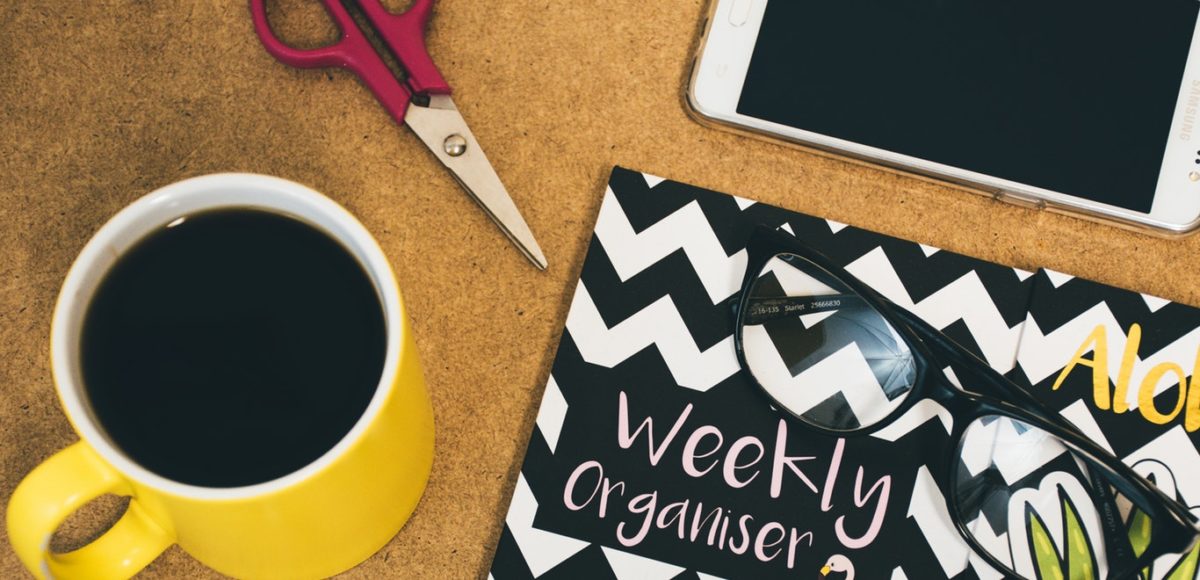 Weekly organizer sits on a desk with cup of black coffee, iPhone, scissors, and reading glasses