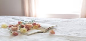 Roses and a book on a bed with a white comforter and sheer curtains blurred in background