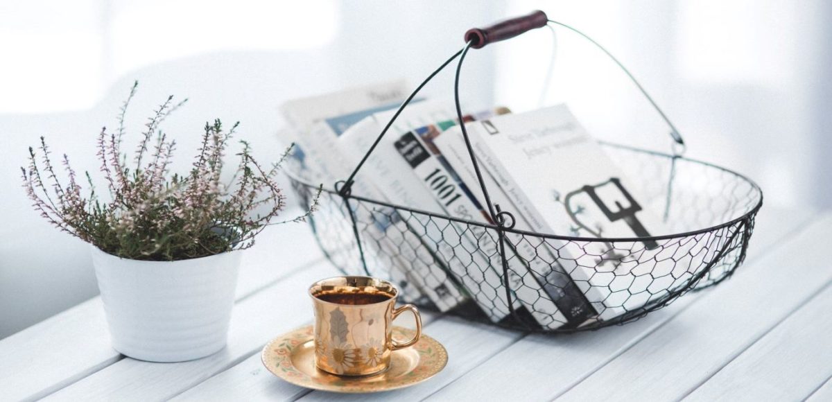 Pretty wire basket with books sitting next to a plant and a brass teacup and saucer on a white table