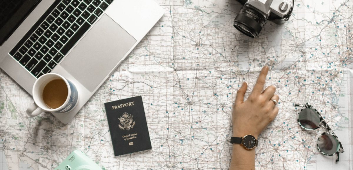 Finger pointing to U.S. map with passport, laptop, camera, and sunglasses on top