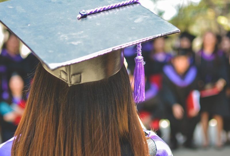 A female graduate in a purple cap and gown faces a large group of grads in the background