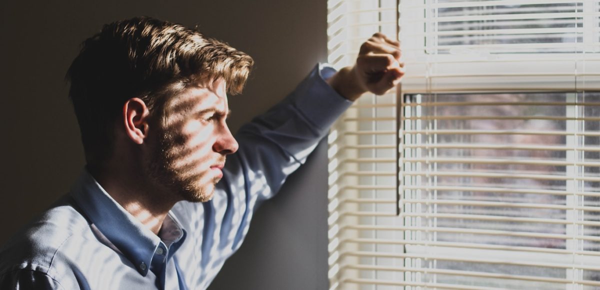 Sad young man looks through the open mini blinds on a window, leaning his arm on the wall