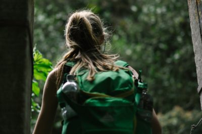 Back view of traveling blonde college female wearing green backpack looking outside an open door