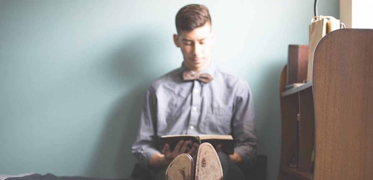 Male college student, smartly dressed, sitting on dorm room bed against a blank wall reading a book