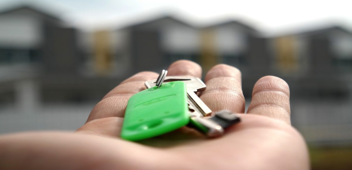 A large hand holding a green house key with blurred home exteriors in the background