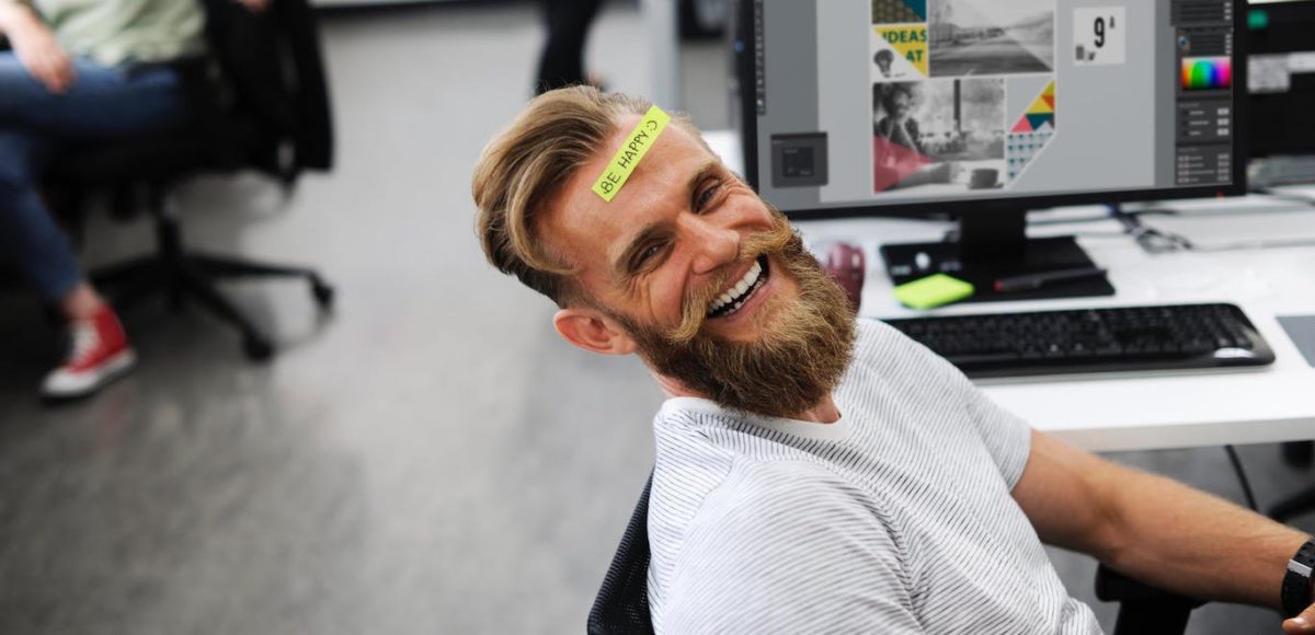 Smiling man with a beard sits in office chair with a yellow note on forehead that says, "Be happy."