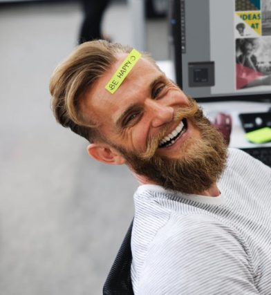 Smiling man with a beard sits in office chair with a yellow note on forehead that says, 
