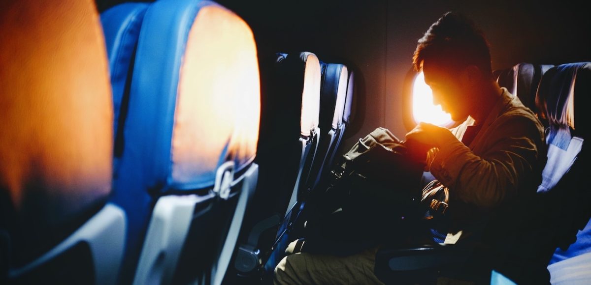 Man sitting in airline seat with carry-on luggage and sunset visible through window