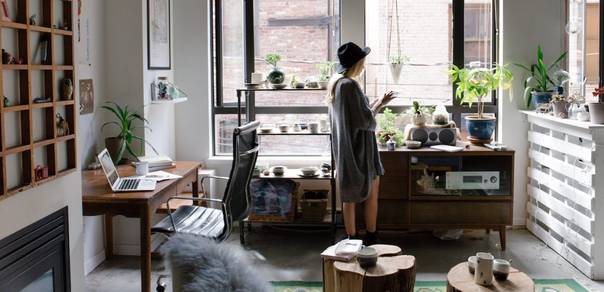 Stylish young woman stands next to a window in a trendy living room filled with plants and a home office