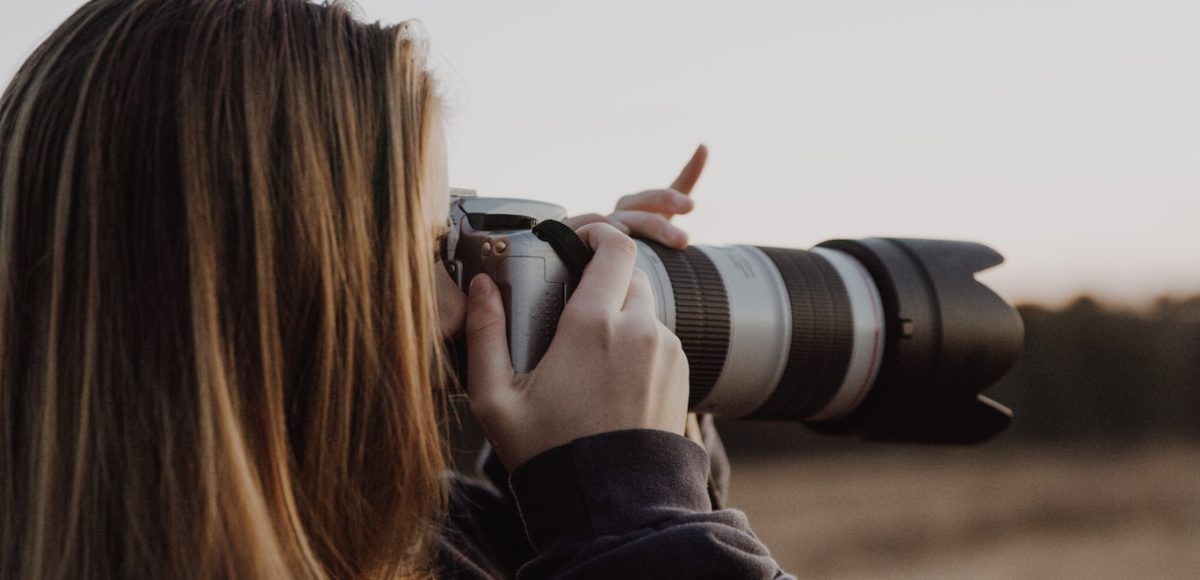 Brunette woman holds DSLR camera with a long lens up to her eyes and adjusts the lens with her hands
