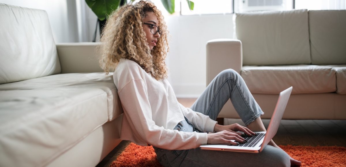 Young woman with curly hair works from home on a laptop from the floor of her living room, leaning against a white sofa in daytime