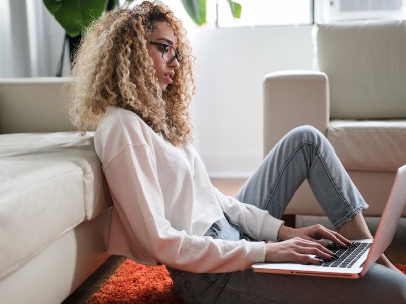 Young woman with curly hair works from home on a laptop from the floor of her living room, leaning against a white sofa in daytime
