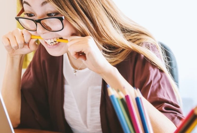 frustrated girl with glasses sitting at desk looking at computer while biting down on a pencil.