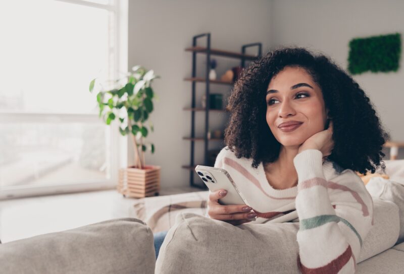 Woman sitting on couch with phone wondering if apartments come furnished in stylish loving room decorated by CORT Furniture Rental.