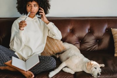 Pensive woman sitting on leather sofa with dog with notebook in lap.