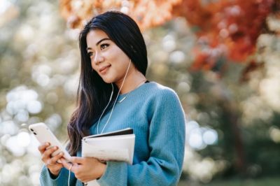 Smiling woman standing in park holding notebook with headphones in ears