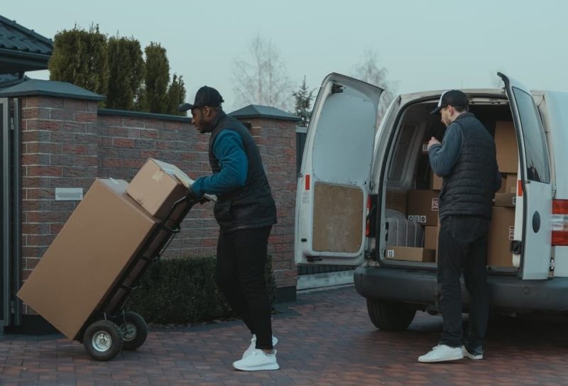 Two movers removing boxes from van and taking them into house on trolley.