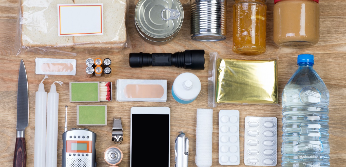 Items on a table that belong in an emergency kit, including: a sandwich, cans of food, bandaids, knife, candles, matches, walkie talkie, phone, medicine, water, batteries, a flashlight, and an emergency blanket