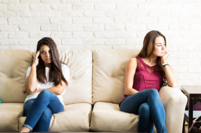 Two women roommates sit next to one another after an argument with distance between them looking sad and angry