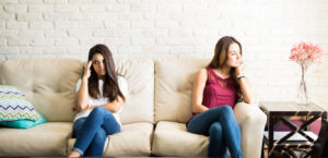 Two women sit on the couch with distance between them looking sad and angry