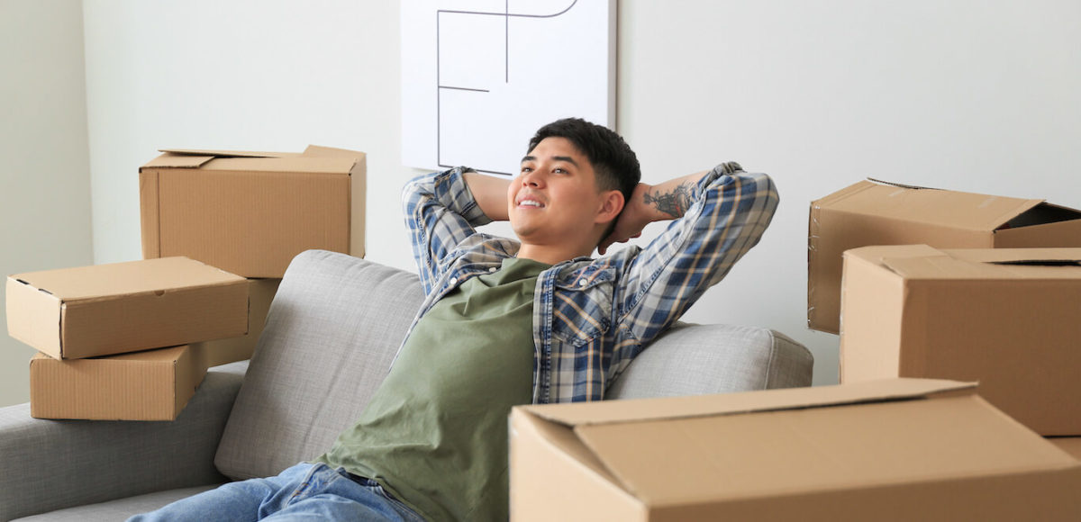 A young man in a flannel relaxes back into a couch surrounded by moving boxes to celebrate moving into a new apartment.