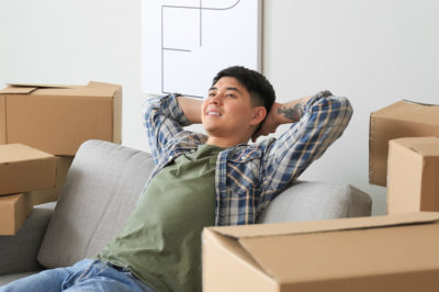 A young man in a flannel relaxes back into a couch surrounded by moving boxes to celebrate moving into a new apartment.
