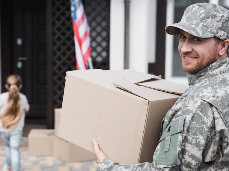 Smiling serviceman carries box to move into new home after military relocation.