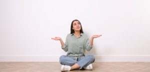 Young woman sitting on floor near white wall indoors after furniture delivery delay.