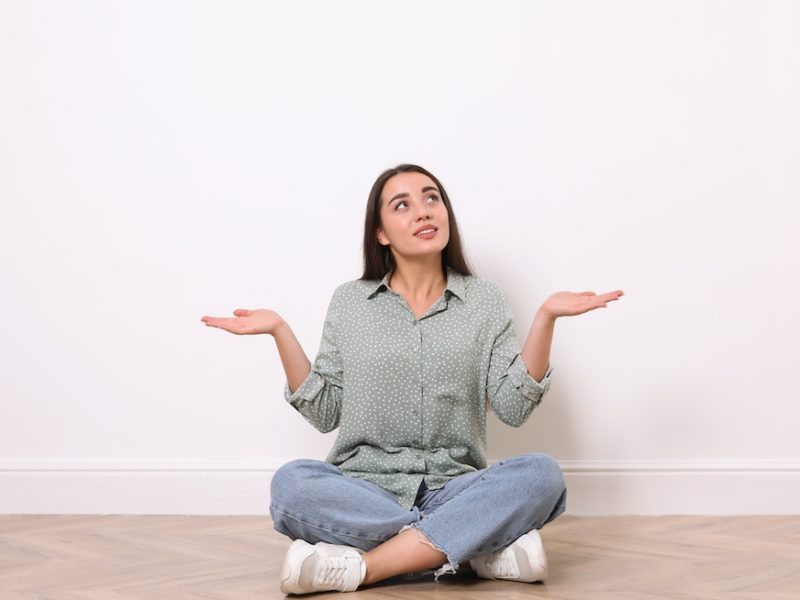 Young woman sitting on floor near white wall indoors after furniture delivery delay.