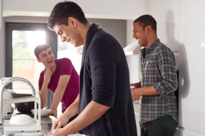 Group Of Male College Students In Shared House Kitchen Washing Up And Hanging Out Together