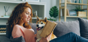 Pretty student young woman is reading book in cozy apartment smiling and petting adorable dog sitting on comfy couch at home. Animals and hobby concept.