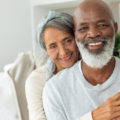 Front view of happy senior diverse couple sitting in a white room in beach house. Authentic Senior Retired Life Concept