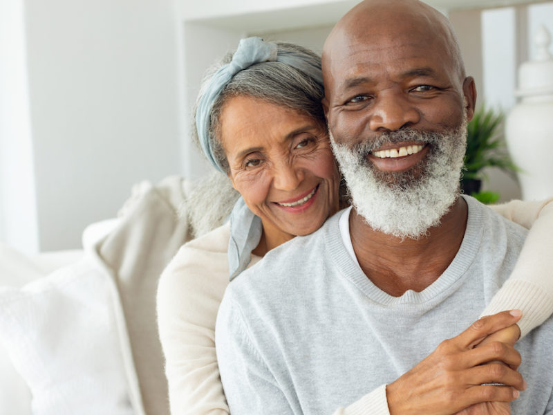 Front view of happy senior diverse couple sitting in a white room in beach house. Authentic Senior Retired Life Concept