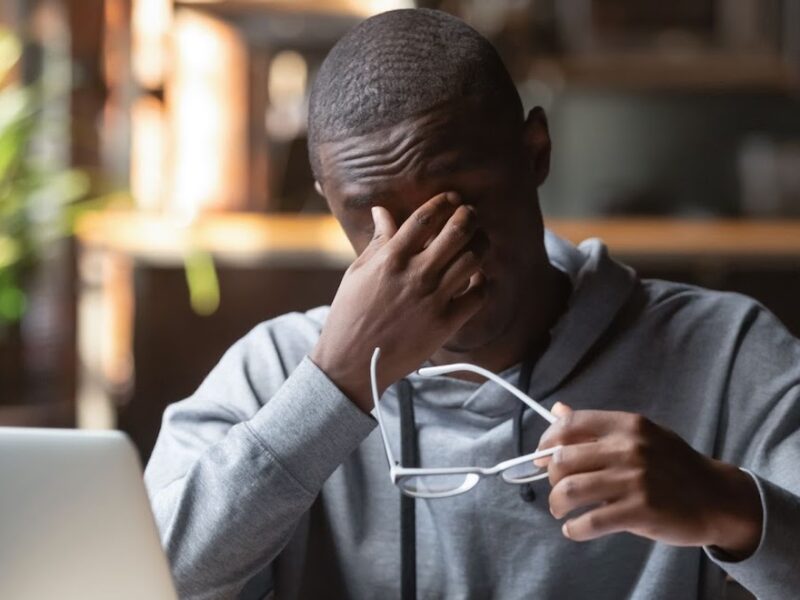 Young black man holding glasses in grey sweatshirt touching forehead to demonstrate exasperation with silver modern laptop in front of him while experiencing a work from home burnout spiral