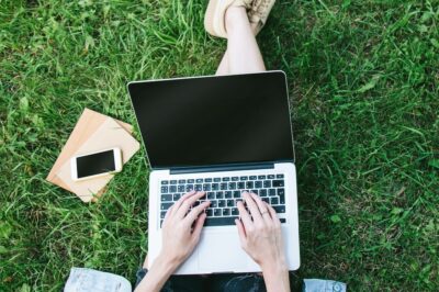 A college student works on a laptop, sitting in green summer grass.