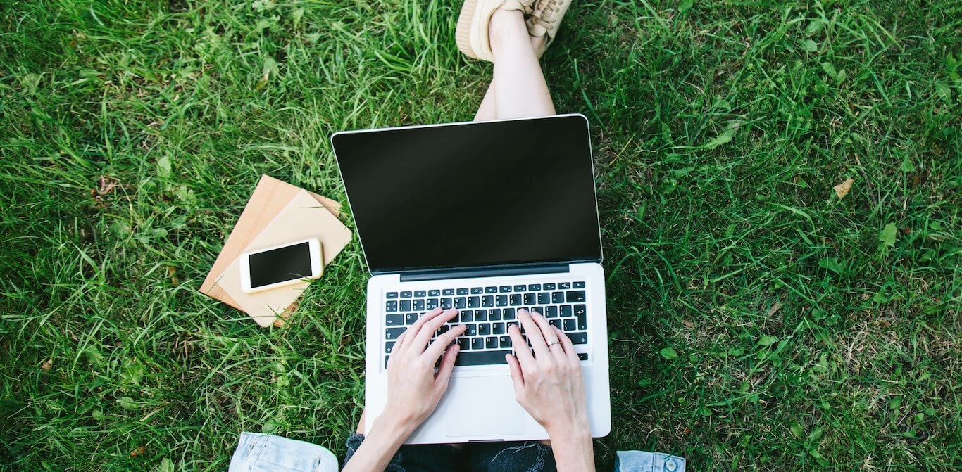 A college student works on a laptop, sitting in green summer grass.