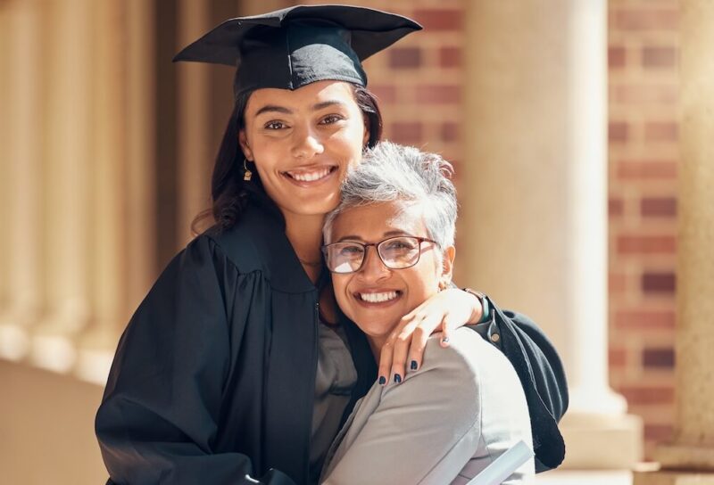 A young woman in a graduation cap and gown hugs her mom.